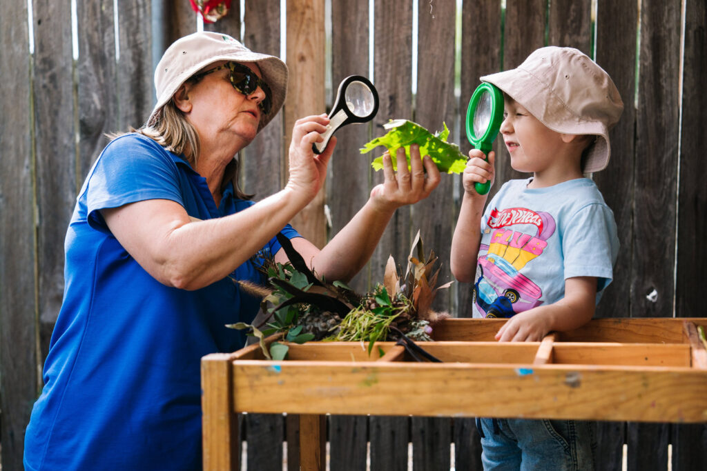 KU Briar Cottage childcare educator and a child looking at a plant with magnifying glasses