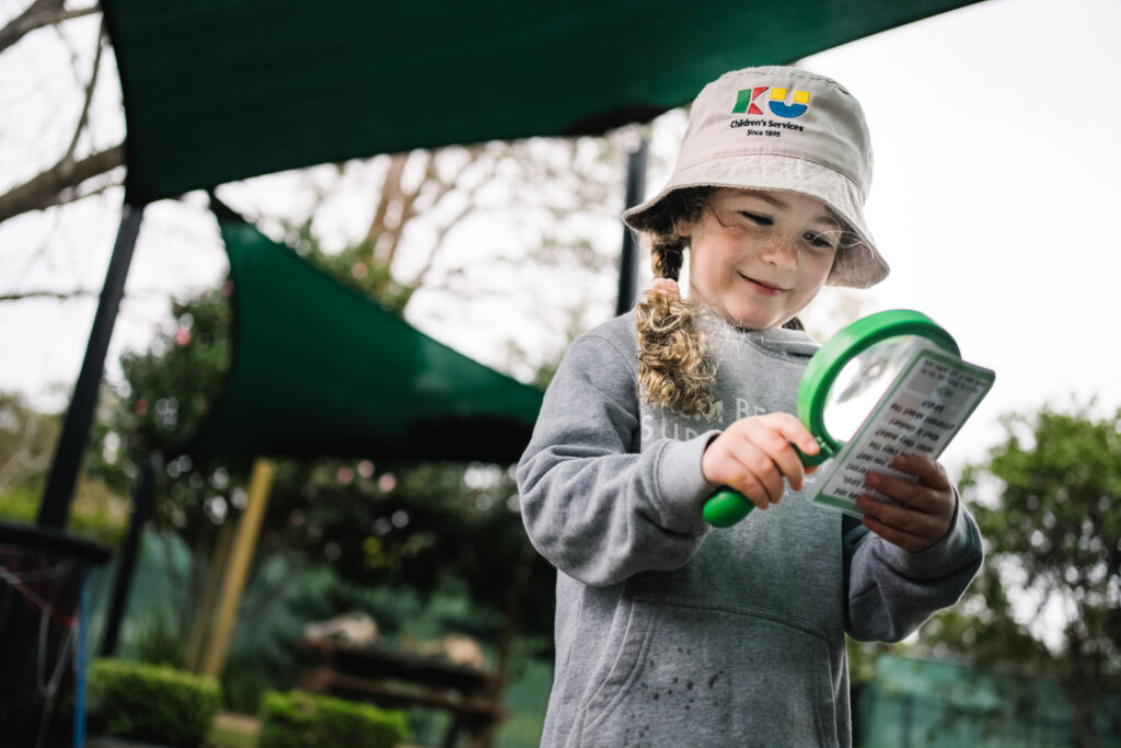 Child using a magnifying glass to study an object at KU Berowra childcare