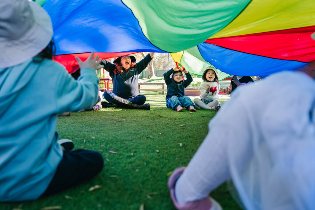 KU Georges Hall childcare educator and children playing with a rainbow parachute