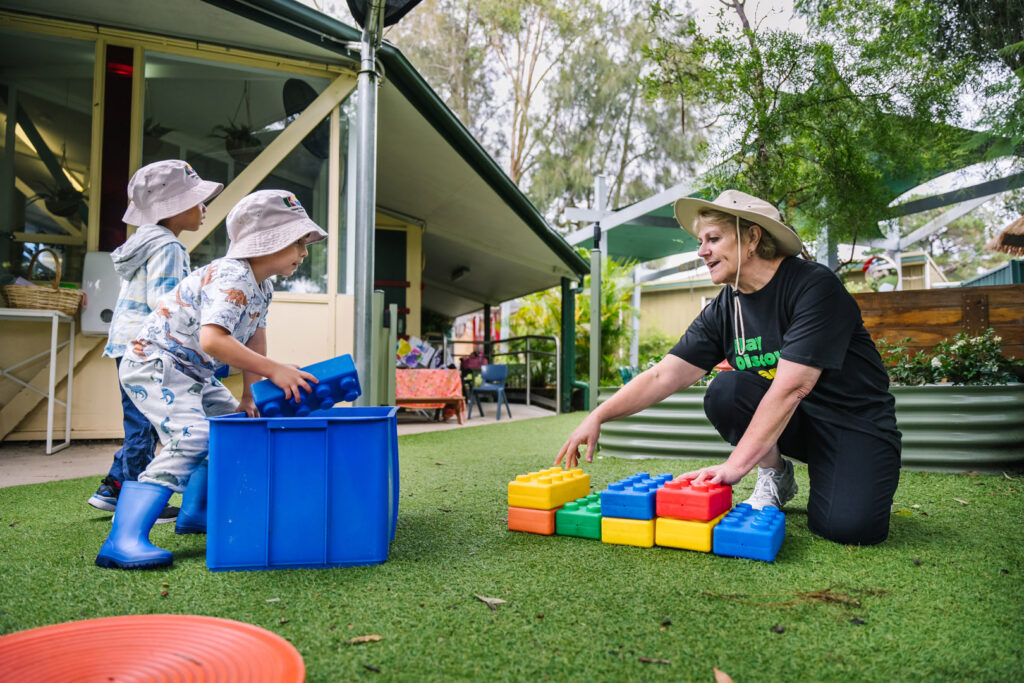 KU Macquarie Fields childcare educator helping children build a block tower outdoors