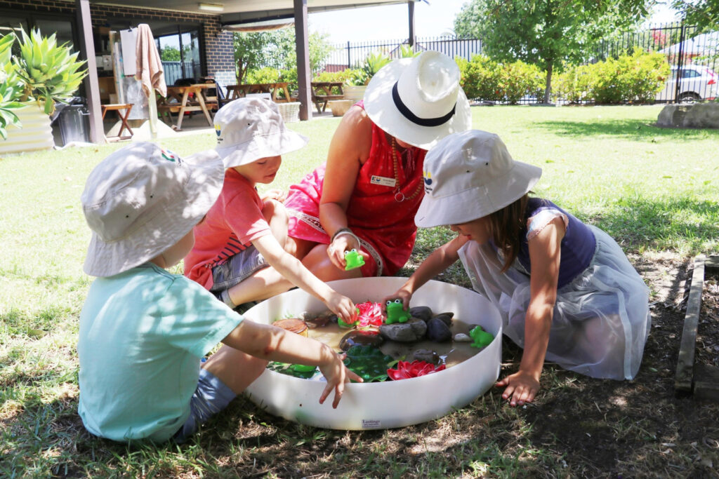 KU Koala childcare educator and children playing with rubber frogs in a pond